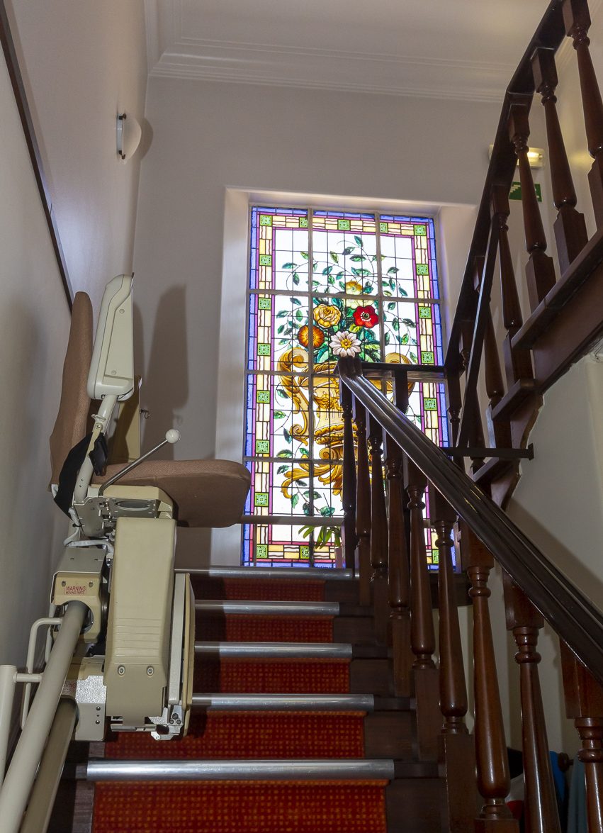 A stairlift beside the staircase below the original stained-glass window